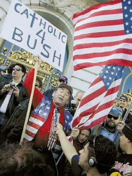 Man in stars-and-stripes vest holding American flag and sign reading "Catholics 4 Bush"