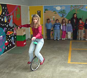 [photo of Danielle Davis riding a unicycle, children watching]