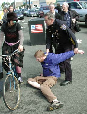 Protester on ground with policeman holding his arm, bicyclist looking on.
