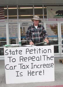 [Petitioner at table in front of store, with sign saying "State petition to repeal the car tax increase is here!"]