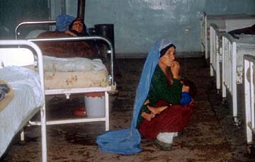 Patient in hospital, surrounded by five other beds, and woman squatting on floor, holding her baby