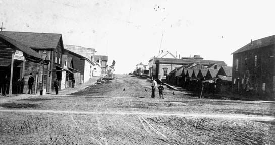 Historic photo depicting unpaved intersection with stores, pedestrians and chickens