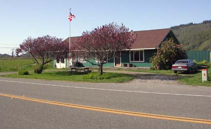 [photo of front of Redwood Lodge Co. main building, with flowering trees, flag pole, picnic table and car in driveway]