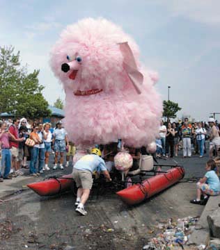 [photo of giant pink poodle kinetic sculpture with floats and crew pushing, spectators watching]