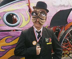 Flatmo wearing suit and mask, standing in front of kinetic sculpture