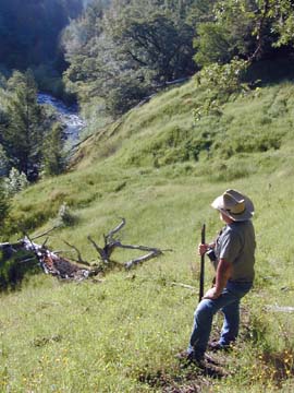 Rodoni standing with his gun on hillside with view down to river