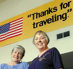Barbara Branco and Fran Tanner of University Travel, standing in front of American flag banner that reads "Thanks for traveling"
