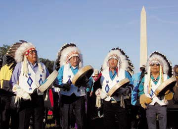 [Four men in feather headdresses, playing drums and singing]