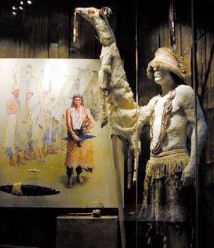[museum exhibit behind glass of manekin wearing white deerskin dance costume and drawing of the dance]