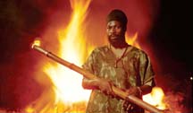 [Capleton holding burning pole, in front of fire, wearing camoflage clothing]
