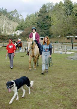 Sally riding a horse on ranch with people and dog following